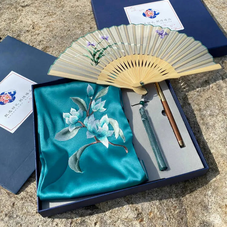 Gift set with embroidered turquoise fabric, wooden fan, and two pens on a stone surface.