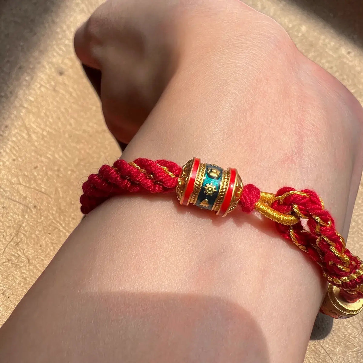 A close-up of a red braided cord bracelet worn on a person's wrist, featuring a decorative cylindrical charm with gold, red, and blue details.