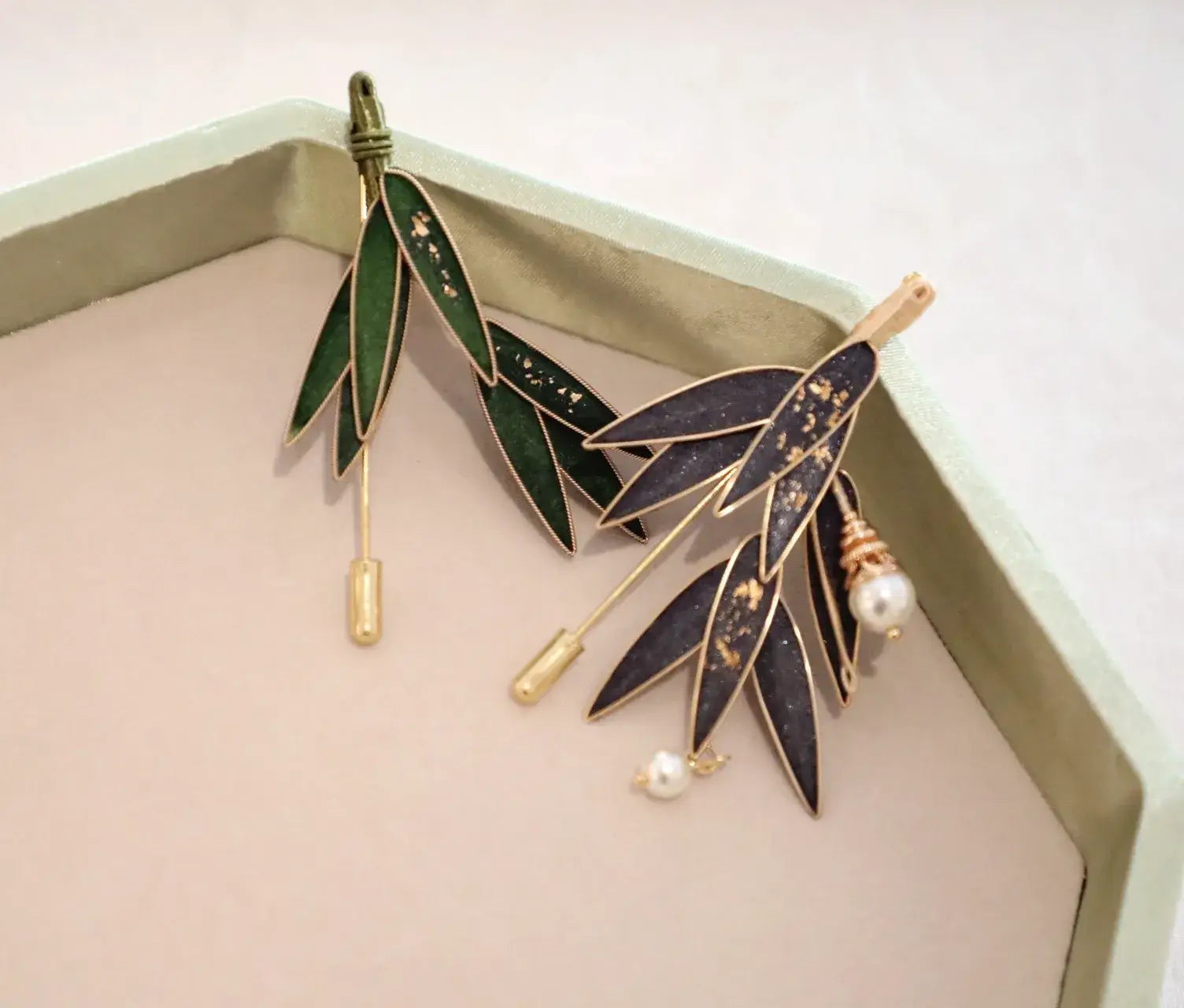 Two leaf-shaped brooches with pearl accents on a light pink surface.