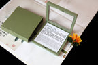 Green jewelry box with a mirror on top, containing text and a flower, placed on a book.