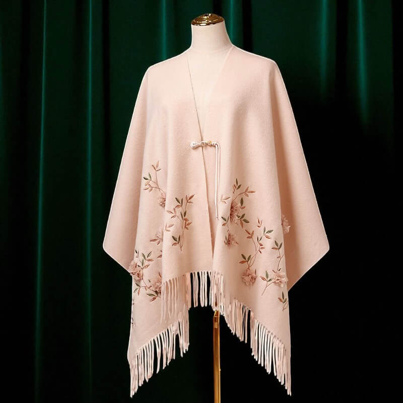 Pink poncho with floral embroidery and fringe on a mannequin against a dark green curtain.