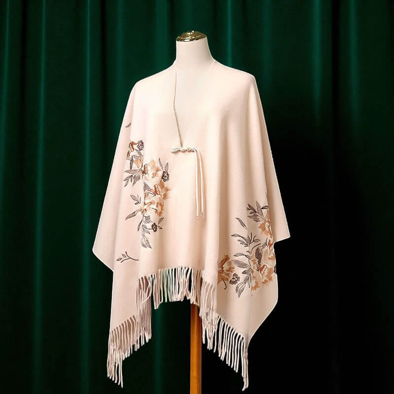 Beige poncho with floral embroidery and fringe on a mannequin against a green curtain background