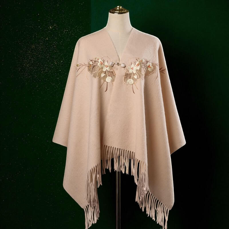 Beige poncho with floral embroidery and fringe on a mannequin against a dark background