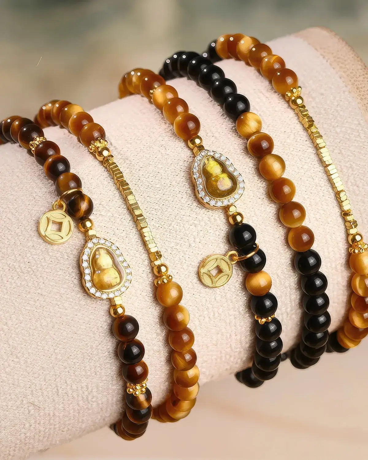Four beaded bracelets displayed on a soft cream fabric wristband. The bracelets feature round beads in brown, black, and tiger-eye patterns, accented with gold-toned chain links, small gold coins, and oval medallions with carved Buddha figures surrounded by clear rhinestones.