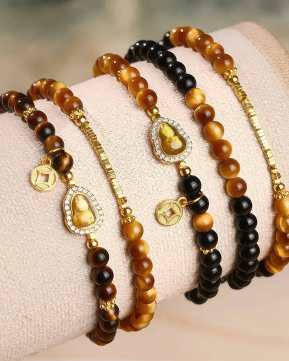 Four beaded bracelets displayed on a soft cream fabric wristband. The bracelets feature round beads in brown, black, and tiger-eye patterns, accented with gold-toned chain links, small gold coins, and oval medallions with carved Buddha figures surrounded by clear rhinestones.