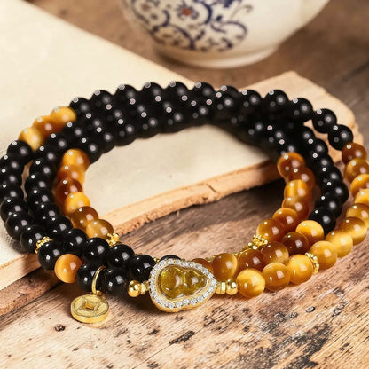 A close-up of a multi-strand beaded bracelet with black, brown, and golden-yellow beads, featuring a gold-tone Buddha charm and a small coin charm, displayed on a rustic wooden surface.