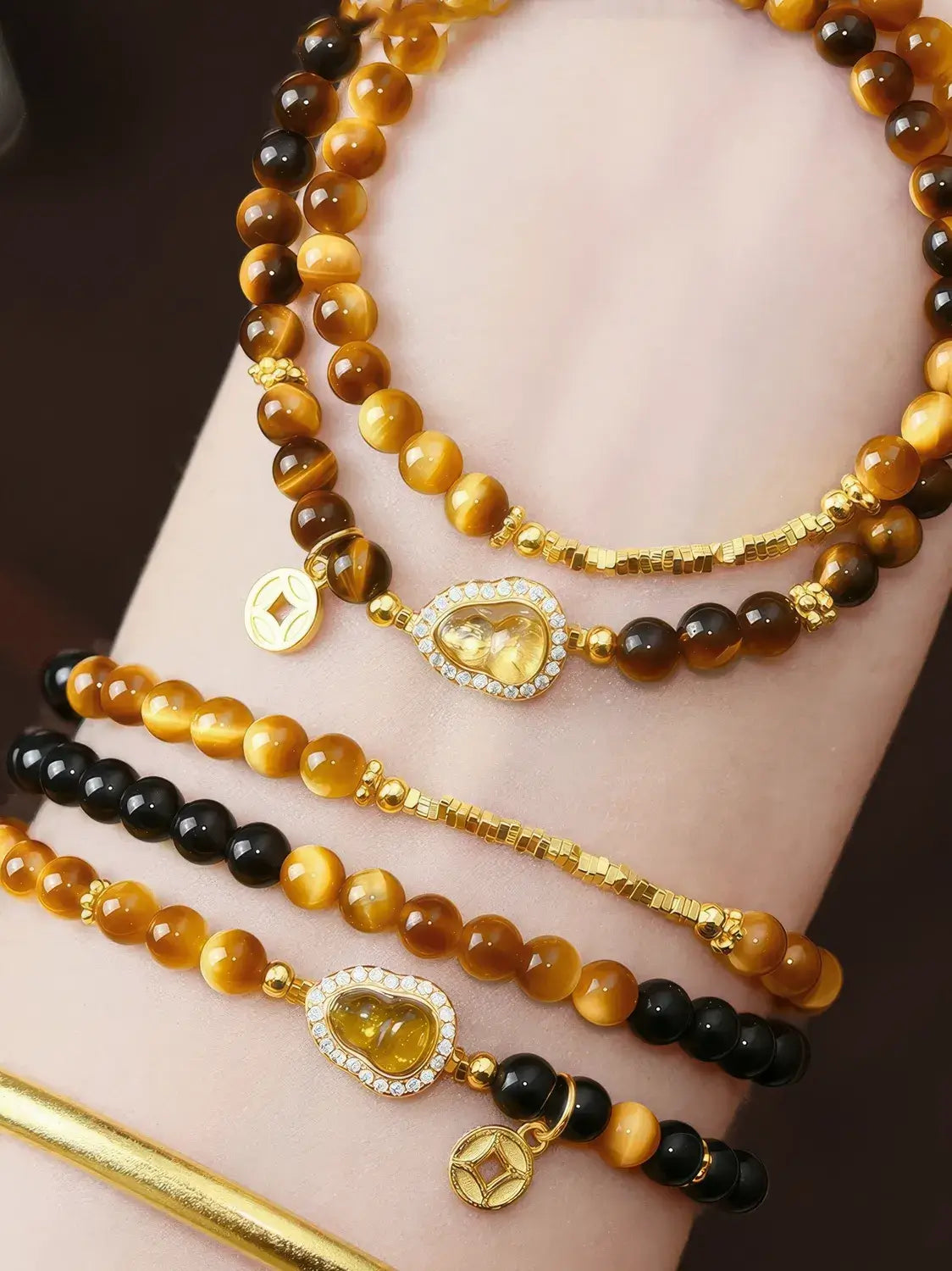 Close-up of a wrist wearing multiple beaded bracelets. The bracelets feature round amber, golden, and black beads with gold-toned charms, including a heart-shaped clear stone charm and small square coin-style charms.