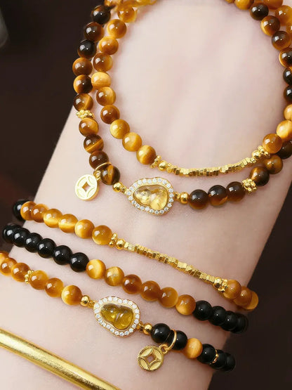 Close-up of a wrist wearing multiple beaded bracelets. The bracelets feature round amber, golden, and black beads with gold-toned charms, including a heart-shaped clear stone charm and small square coin-style charms.