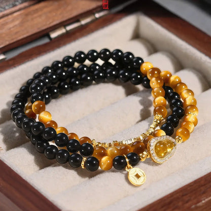A set of three beaded bracelets displayed in a jewelry box. The bracelets feature glossy black beads, golden brown tiger's eye beads, and polished gold-toned metal accents, including a heart-shaped charm with a clear crystal halo and a small gold coin charm.