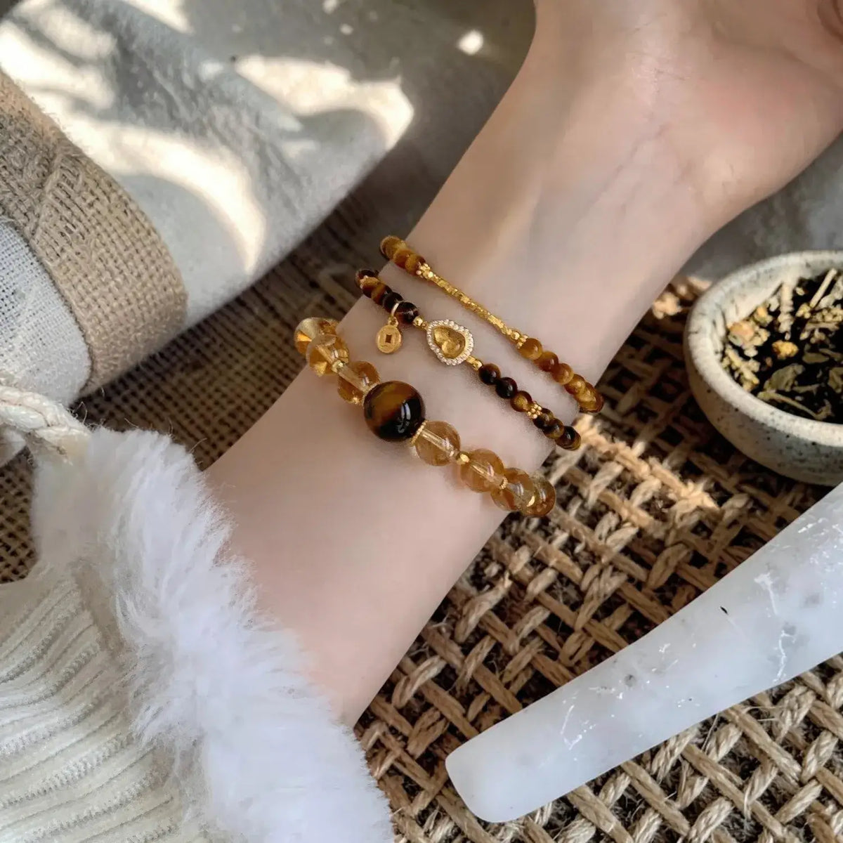 A close-up of a person's wrist wearing three layered beaded bracelets: a gold chain bracelet with a small heart charm, a dark brown and amber bead bracelet, and a translucent yellow-orange stone bead bracelet. The wrist is resting on a woven mat with a white stone cone and a small bowl of dried botanicals nearby.