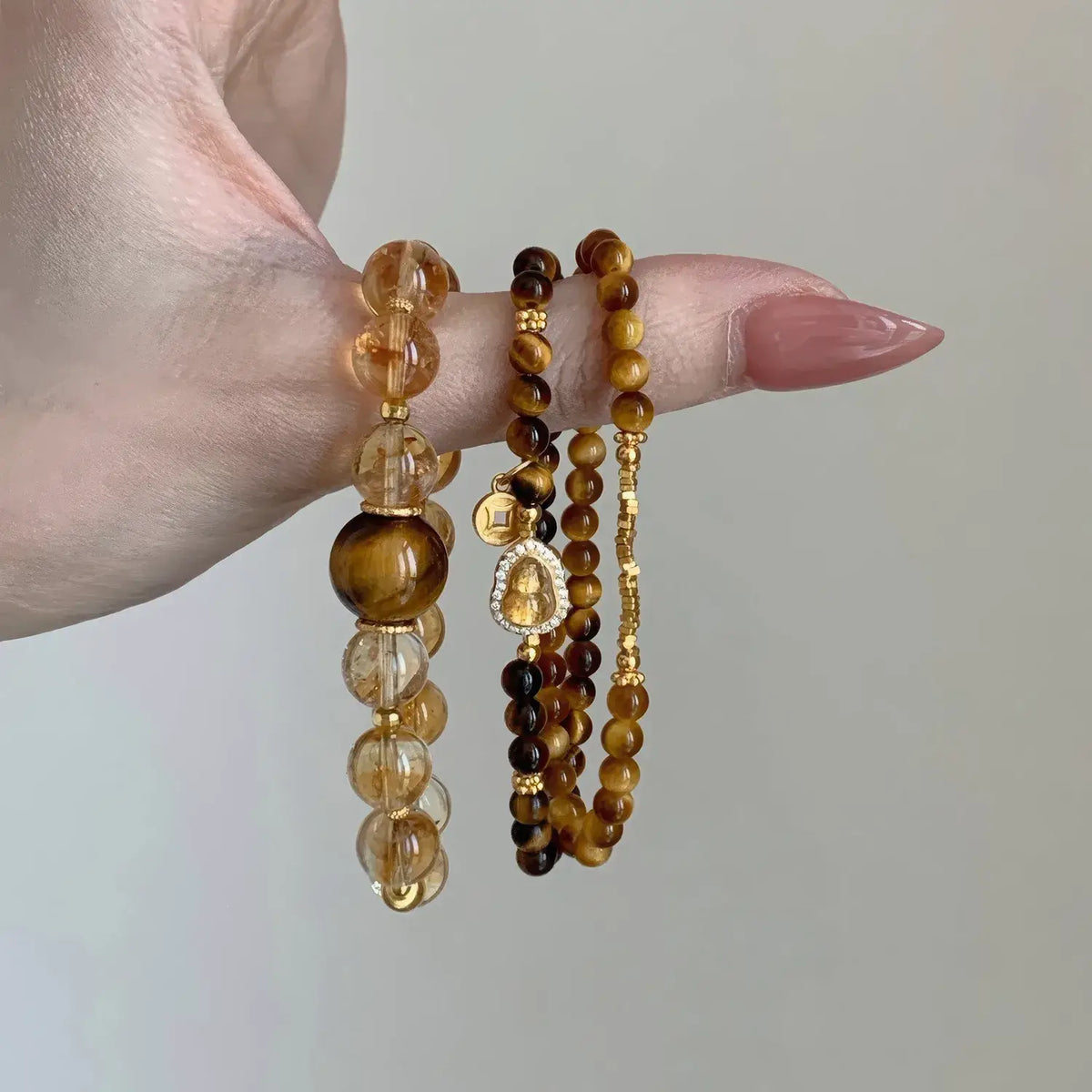 Three beaded stretch bracelets displayed on a person's finger against a plain light gray background. The bracelets include clear faceted beads, brown tiger's eye beads, and gold-toned beaded strands with a small gold charm.