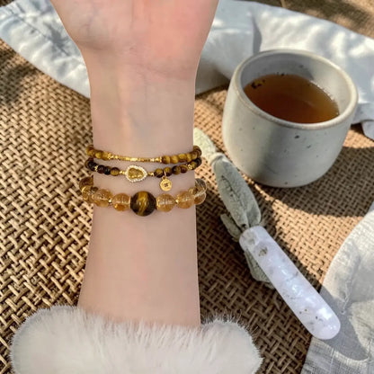 Three beaded bracelets displayed on a person's wrist, featuring yellow tiger's eye, dark brown beads, and gold-toned metal charms, set against a cozy background with a cup of tea and a white stone tool.