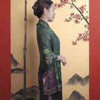 Woman in a traditional green outfit standing against a decorative wall with bamboo and cherry blossom branches.