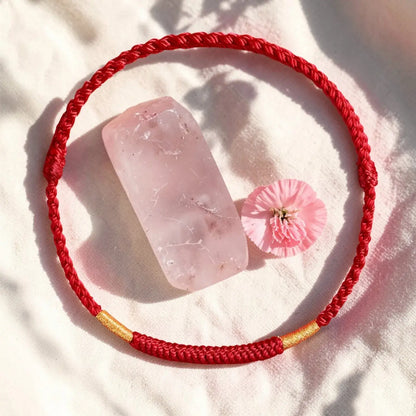Red braided bracelet with gold accent on a white surface with pink crystal and flower.