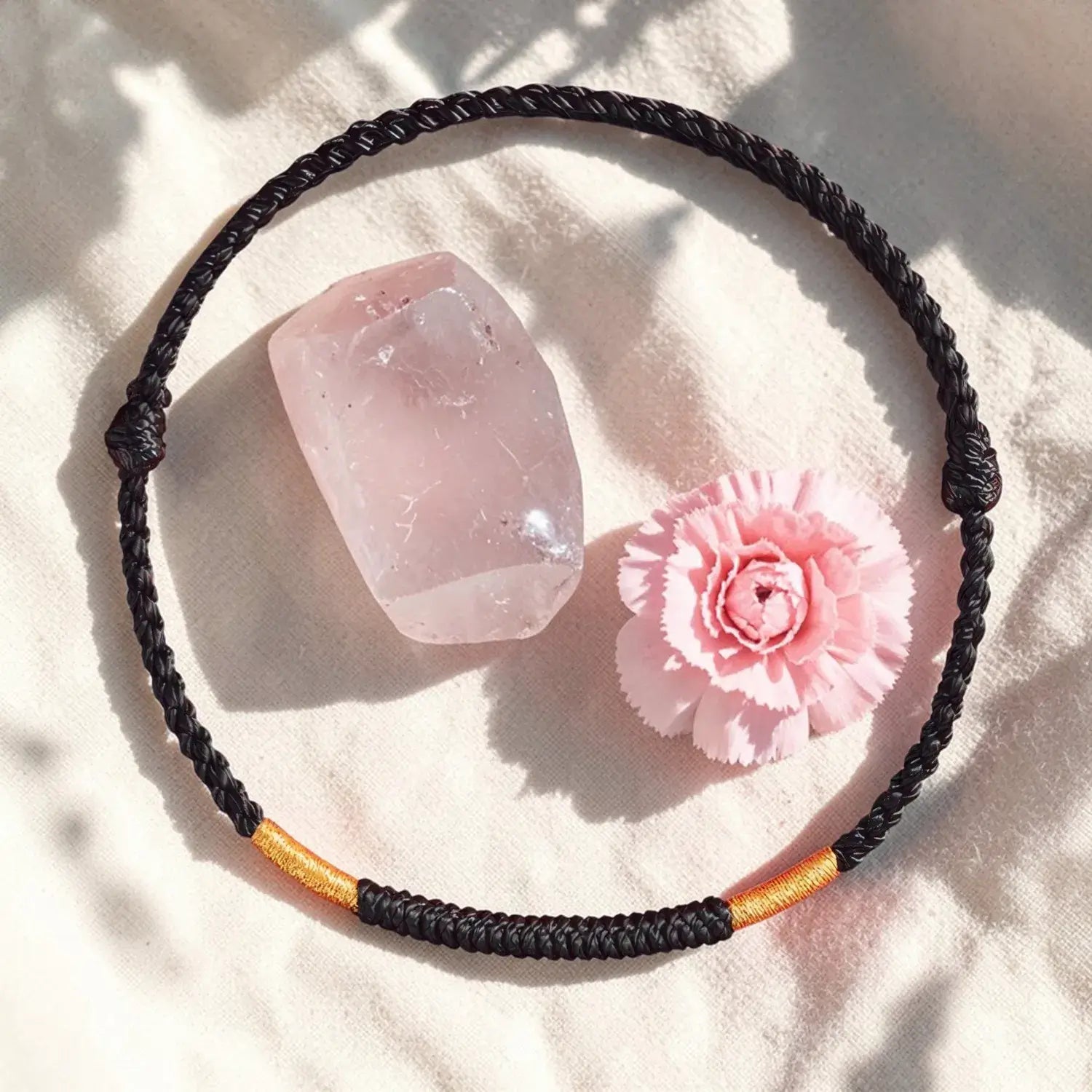 Black braided necklace with gold accent on a light pink surface with a pink crystal and flower.