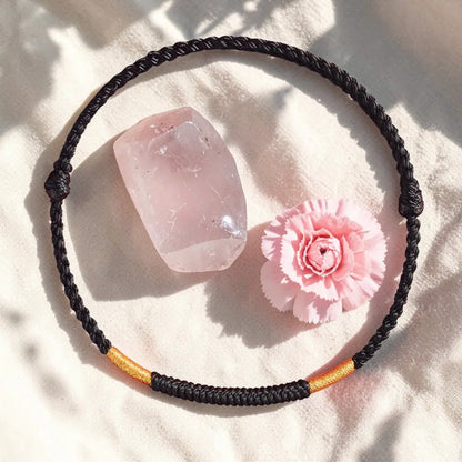 Black braided necklace with gold accent on a light pink surface with a pink crystal and flower.