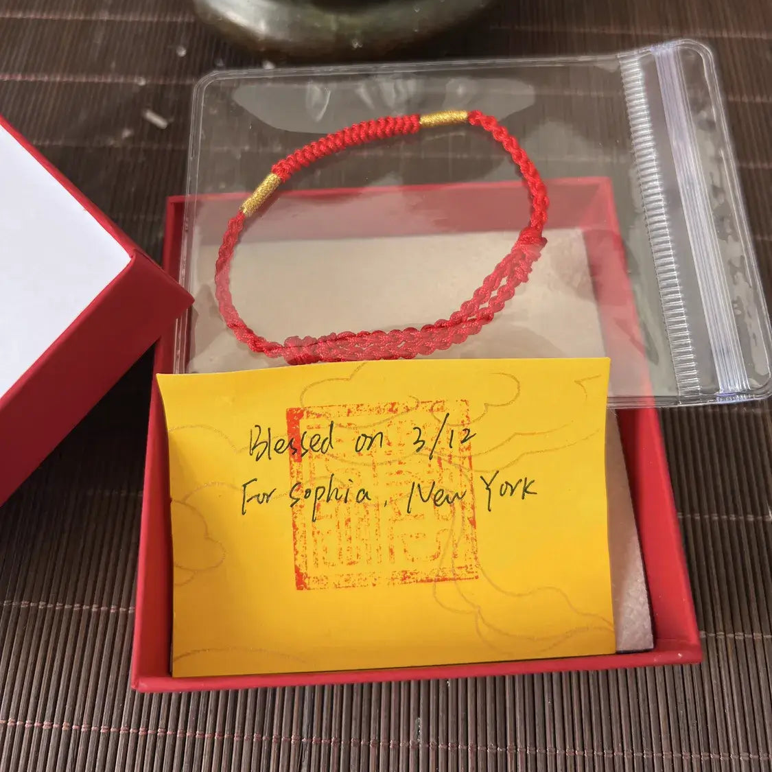 Red beaded bracelet on a card with 'Blessed on 2/13 for Sophia, New York' text, placed on a dark surface with incense and bell in the background.