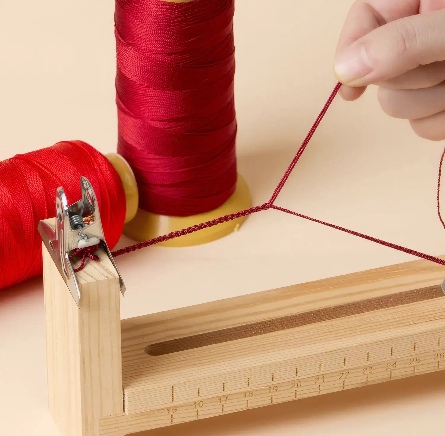 Person threading red thread through a wooden sewing device with spools of red thread in the background.
