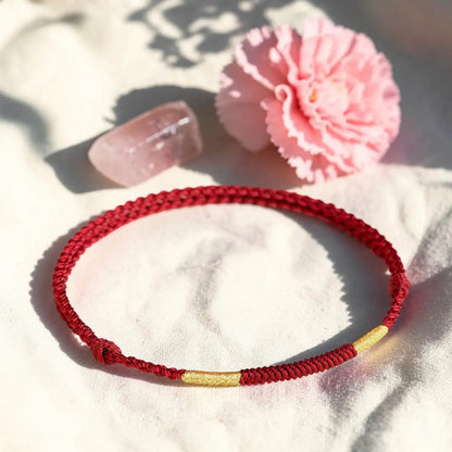 Red braided bracelet with a gold accent on a white surface with a pink flower and clear object.