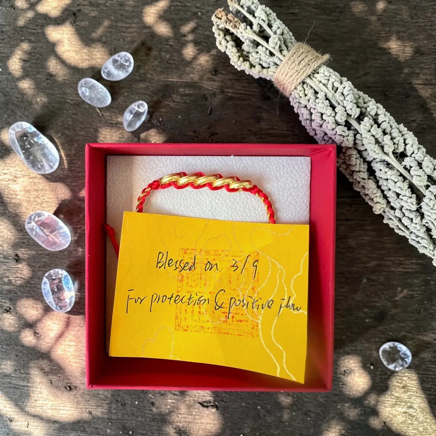 Bracelet with a yellow card in a red box on a wooden surface with crystals and herbs.