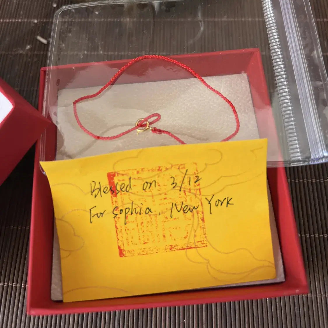 Small red box with a yellow card inside, containing a red string and small object on a textured surface.