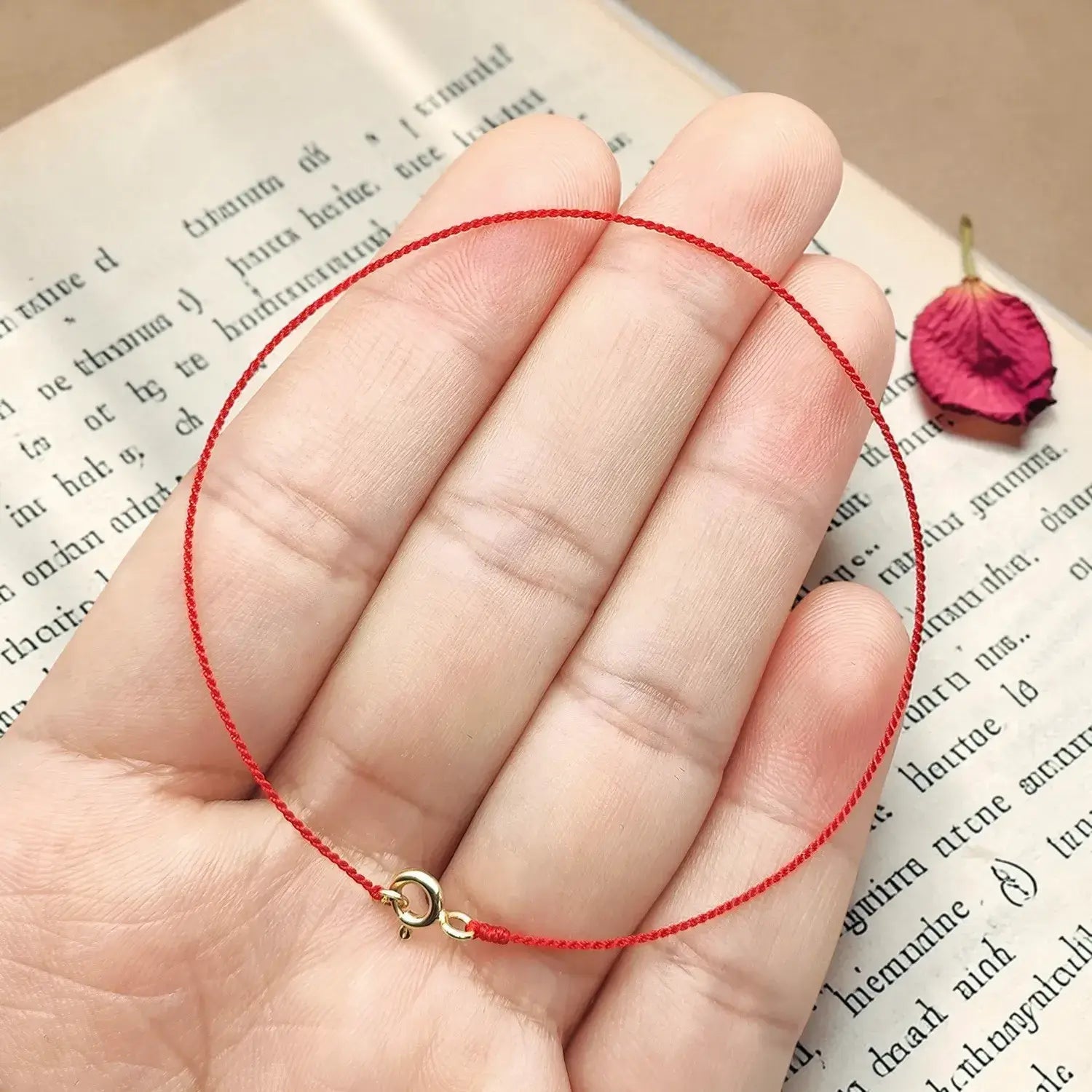 Hand holding a red string bracelet with a gold clasp on an open book with a dried rose petal.