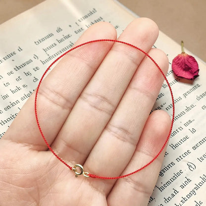 Hand holding a red string bracelet with a gold clasp on an open book with a dried rose petal.